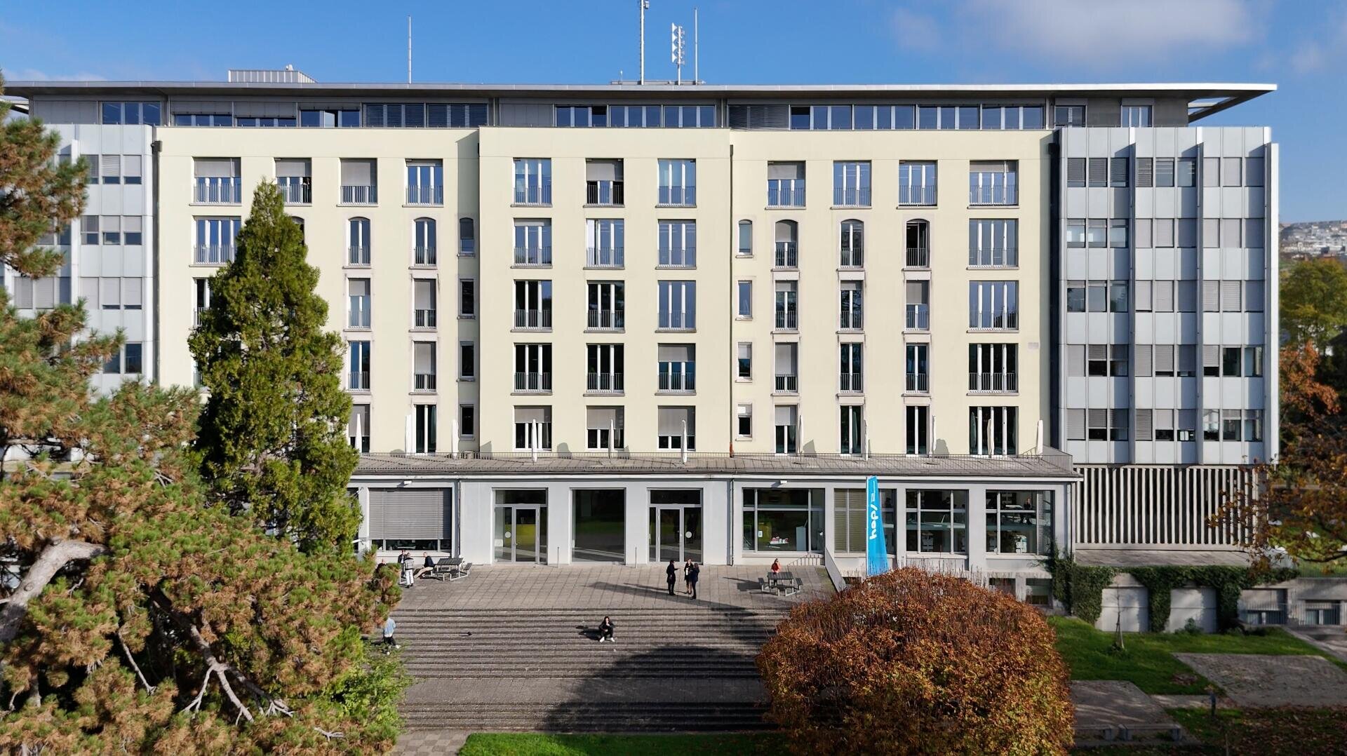 Un immeuble moderne à plusieurs étages, doté de grandes baies vitrées et d'une façade beige, entouré d'arbres et de verdure. Des gens se promènent et sont assis près des marches de l'entrée, sous un ciel bleu.