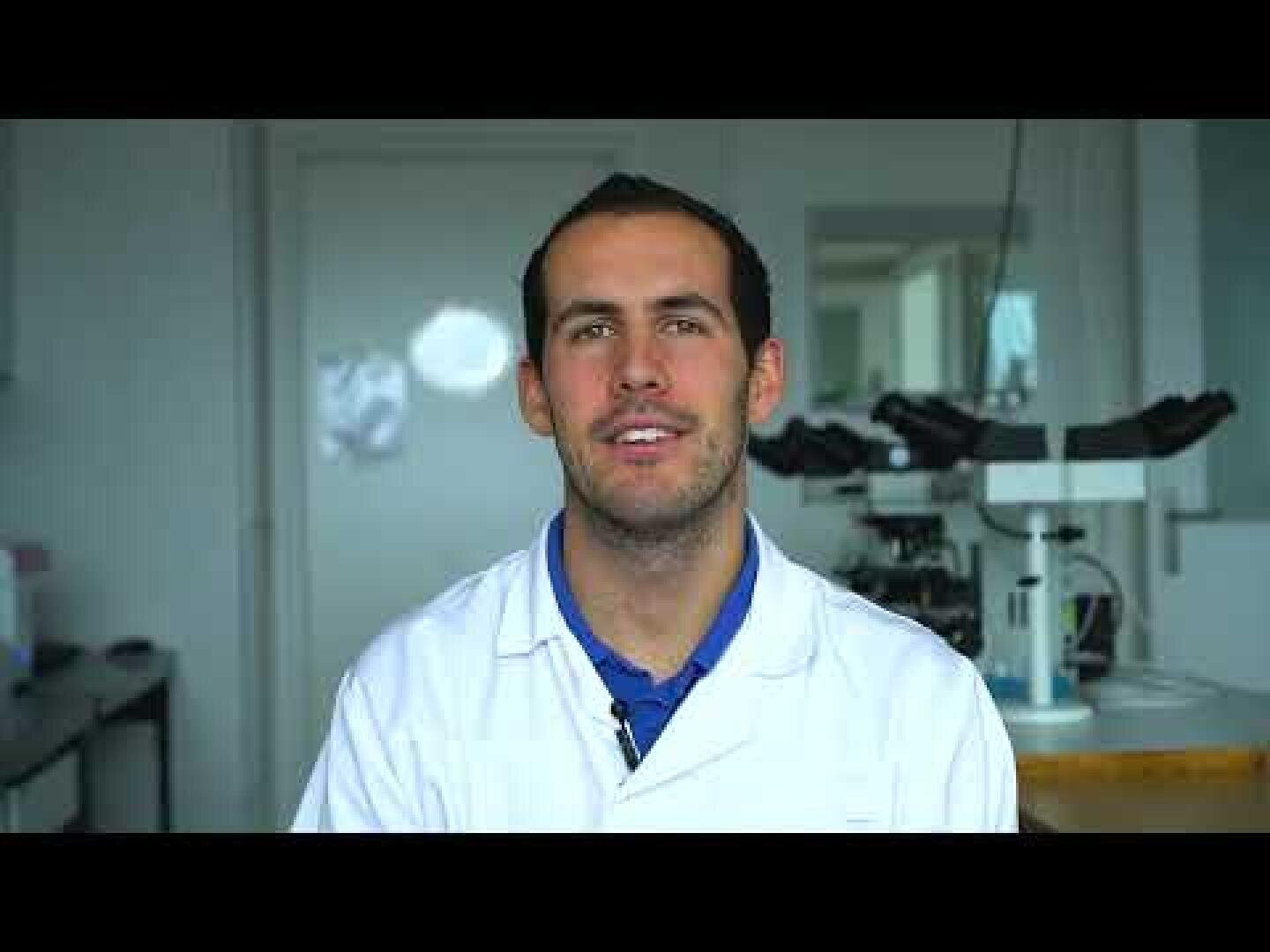 Un homme portant une blouse blanche est assis dans un laboratoire avec du matériel scientifique, dont un microscope, visible à l'arrière-plan. Il fait face à la caméra et sourit légèrement.