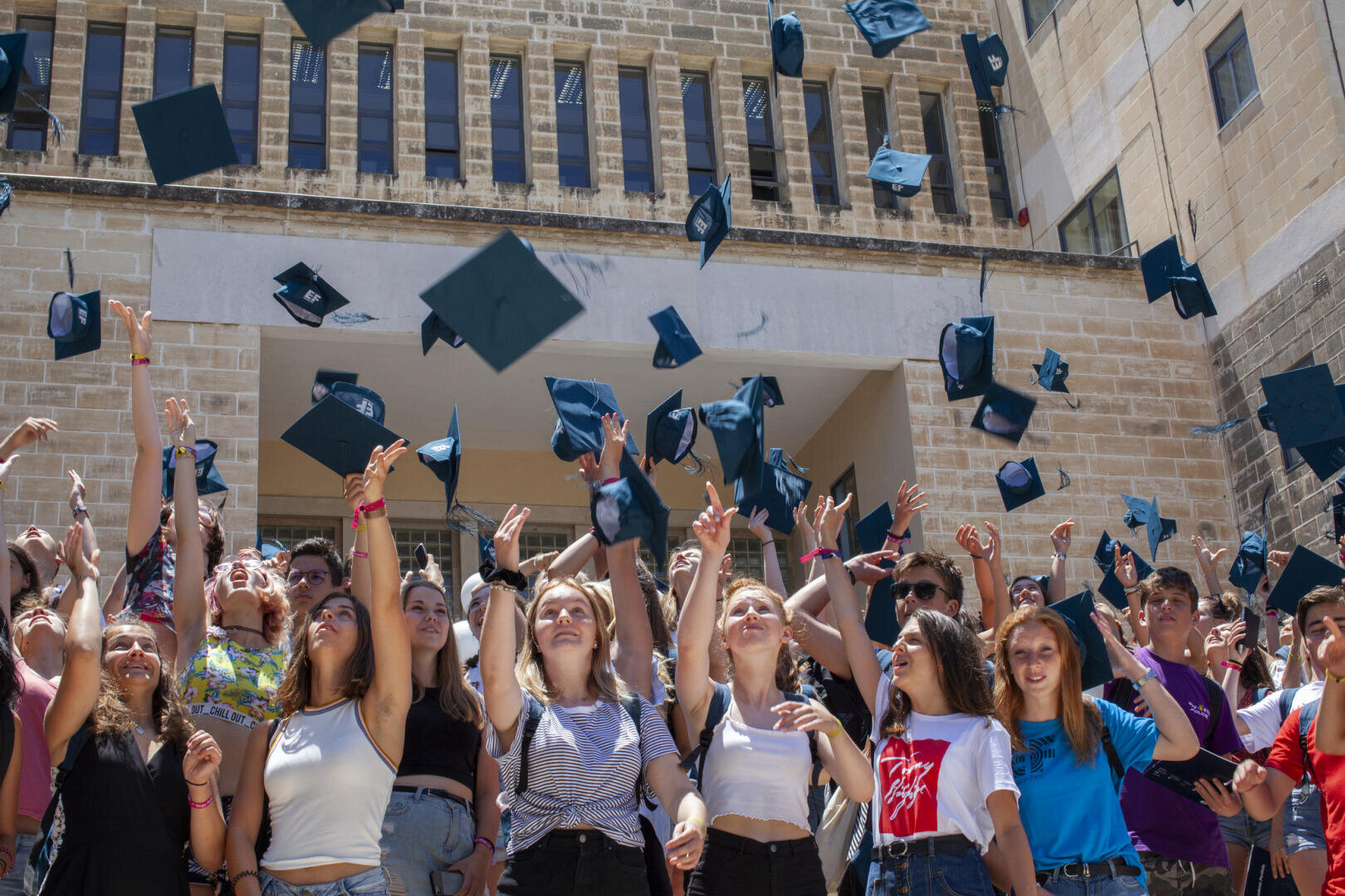 Un grand groupe d'étudiants souriants célèbre la remise des diplômes en plein air, en lançant des casquettes noires en l'air devant un bâtiment en pierre, par une journée ensoleillée.