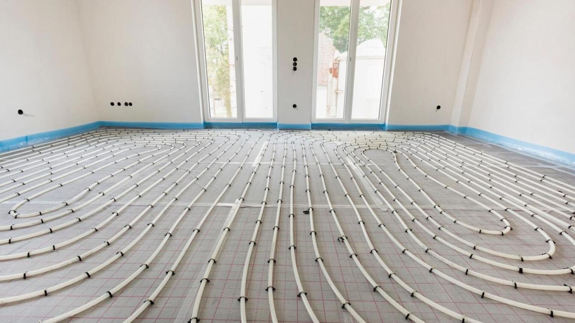 Des tuyaux en plastique blanc pour le chauffage par le sol sont disposés en grille et en serpentin dans une pièce inachevée avec de grandes fenêtres et des murs nus. Une isolation bleue borde les murs de la pièce.