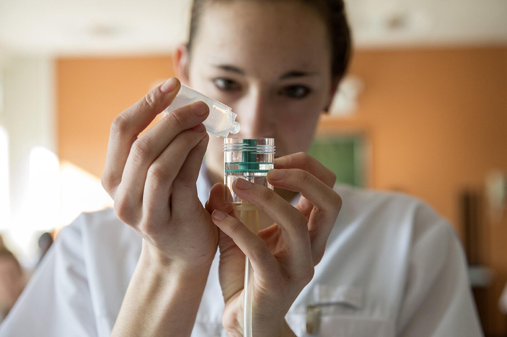 Une personne en blouse blanche verse soigneusement un liquide d'une petite bouteille dans un récipient transparent, concentrée sur sa tâche. L'arrière-plan est légèrement flouté, mettant en valeur la précision du travail.