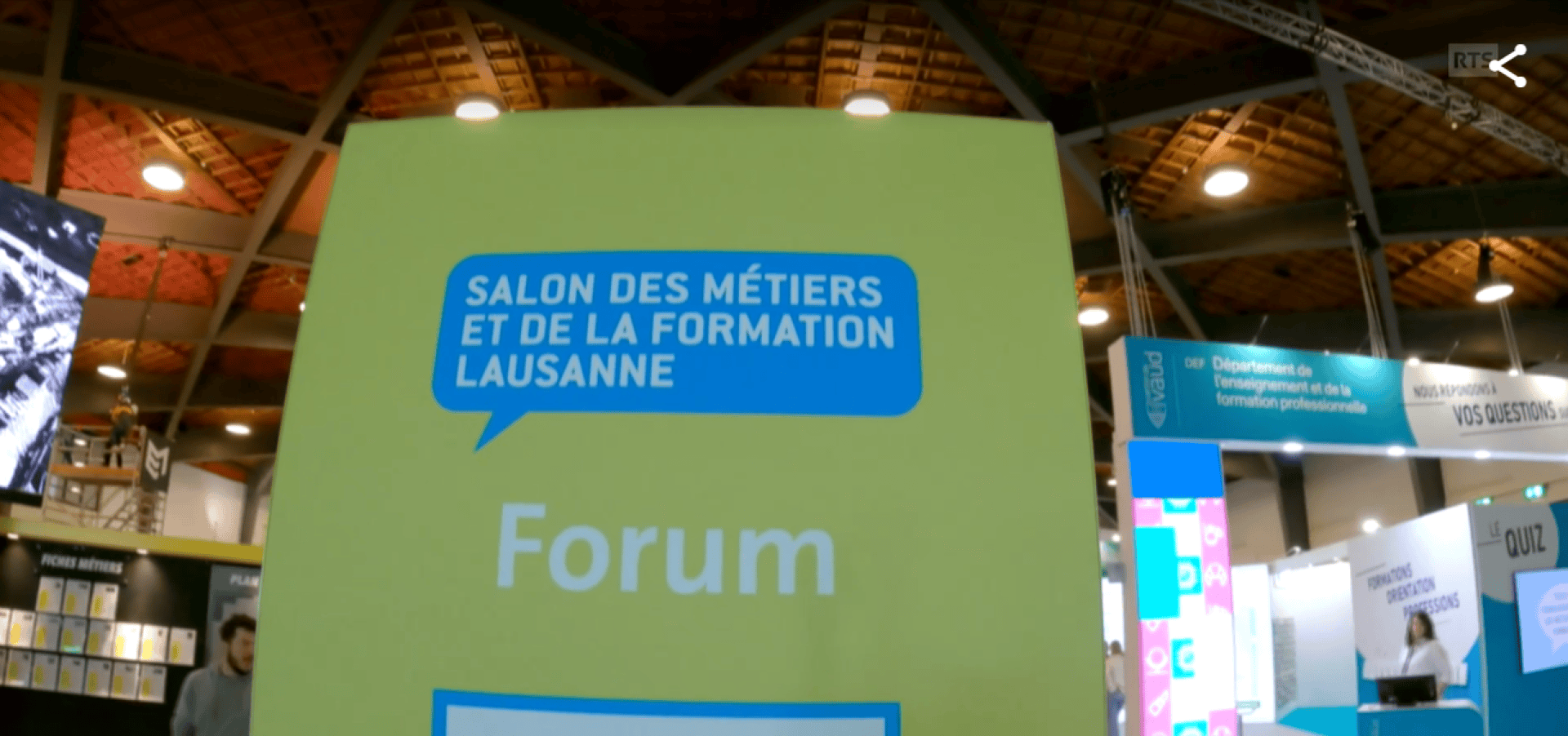 Un grand panneau vert indique Salon des Métiers et de la Formation Lausanne Forum. Des stands d'exposition et des personnes sont visibles à l'arrière-plan sous un plafond en bois.