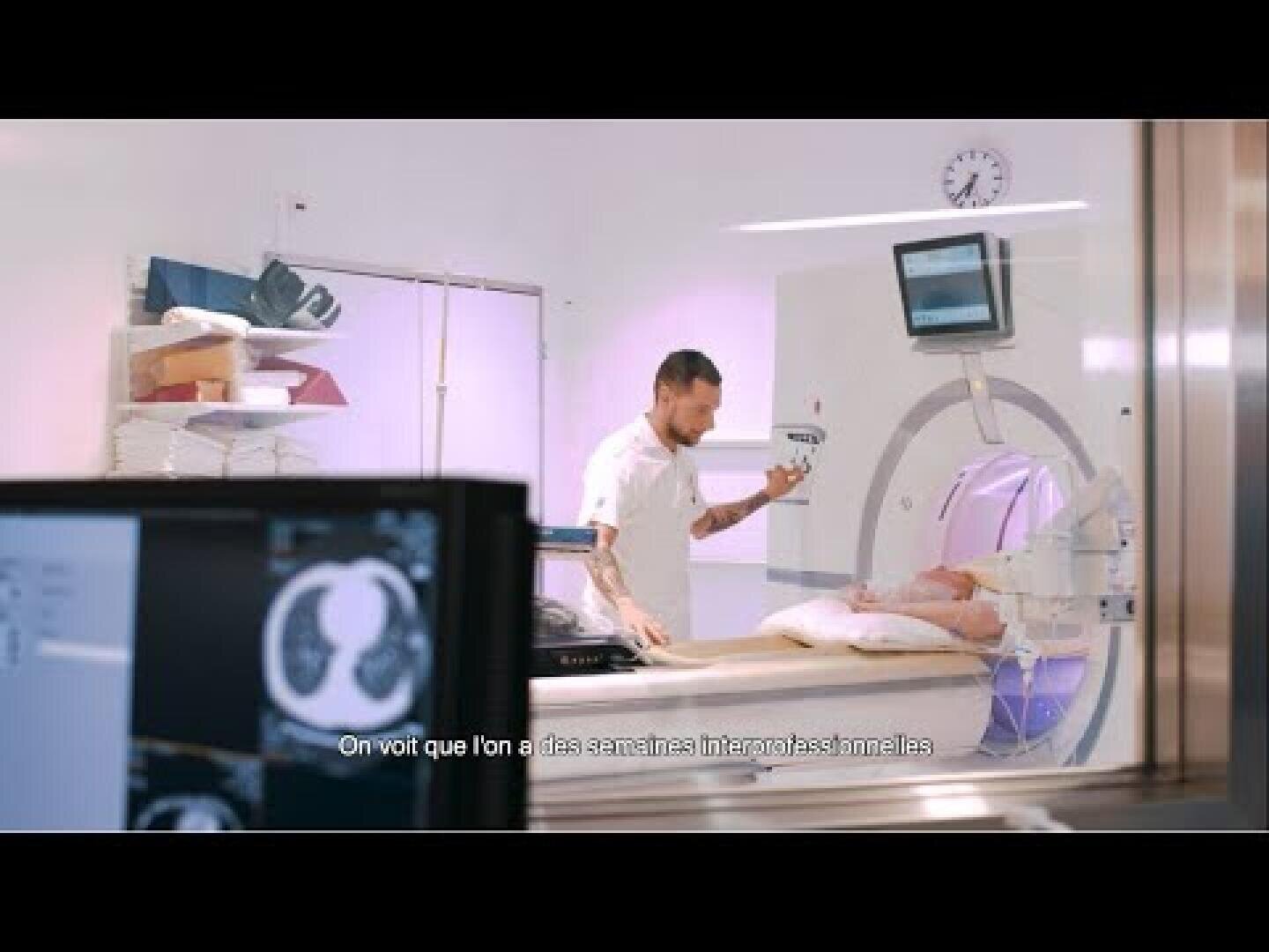 Un professionnel de la santé se tient à côté d'un patient allongé dans un scanner, tenant un appareil. Des moniteurs affichent des images numérisées et des sous-titres en français apparaissent au bas de l'image. La scène est vue à travers une vitre.