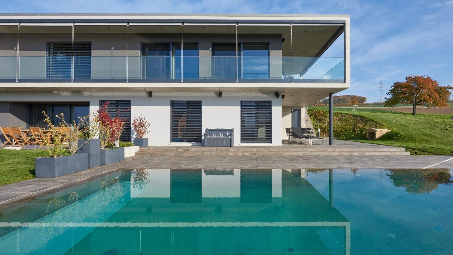 Maison moderne de deux étages avec un grand balcon vitré, des fenêtres allant du sol au plafond et une piscine au premier plan ; entourée d'une pelouse verte et d'un arbre avec des feuilles d'automne à l'arrière-plan.