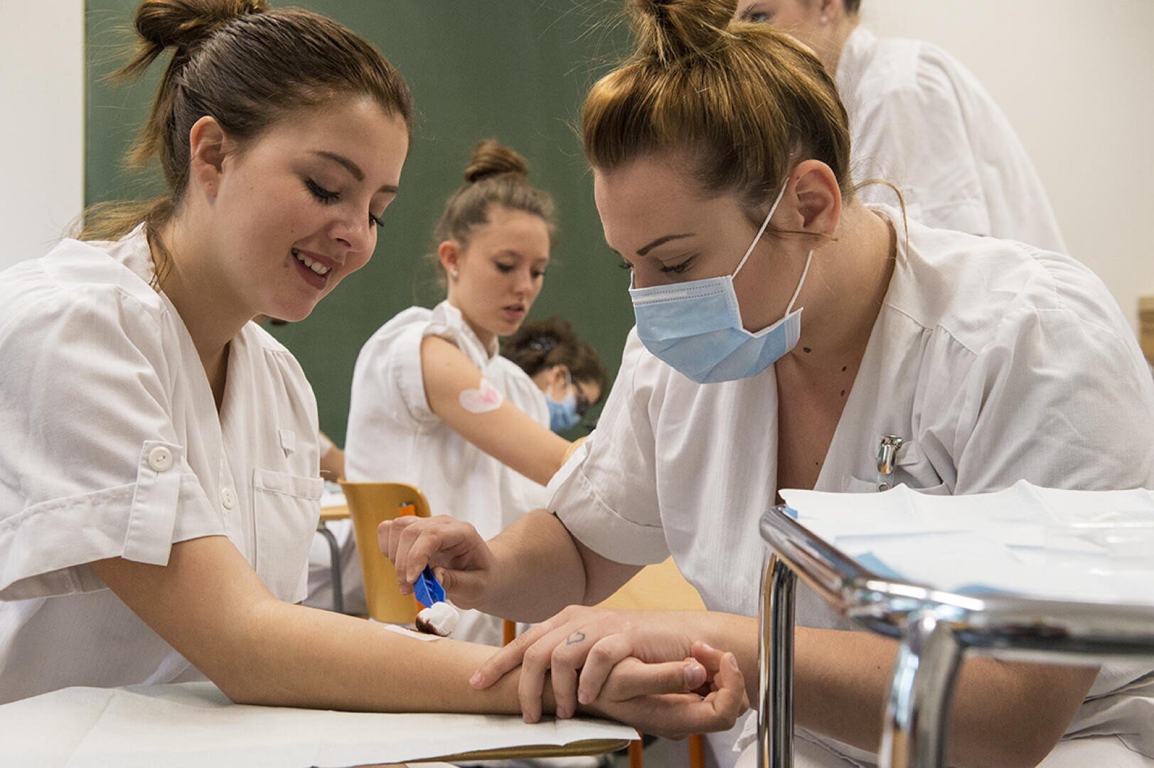 Une infirmière portant un masque se prépare à prélever du sang sur le bras d'une jeune femme souriante, tandis que d'autres femmes en uniforme blanc effectuent des procédures médicales similaires à l'arrière-plan.