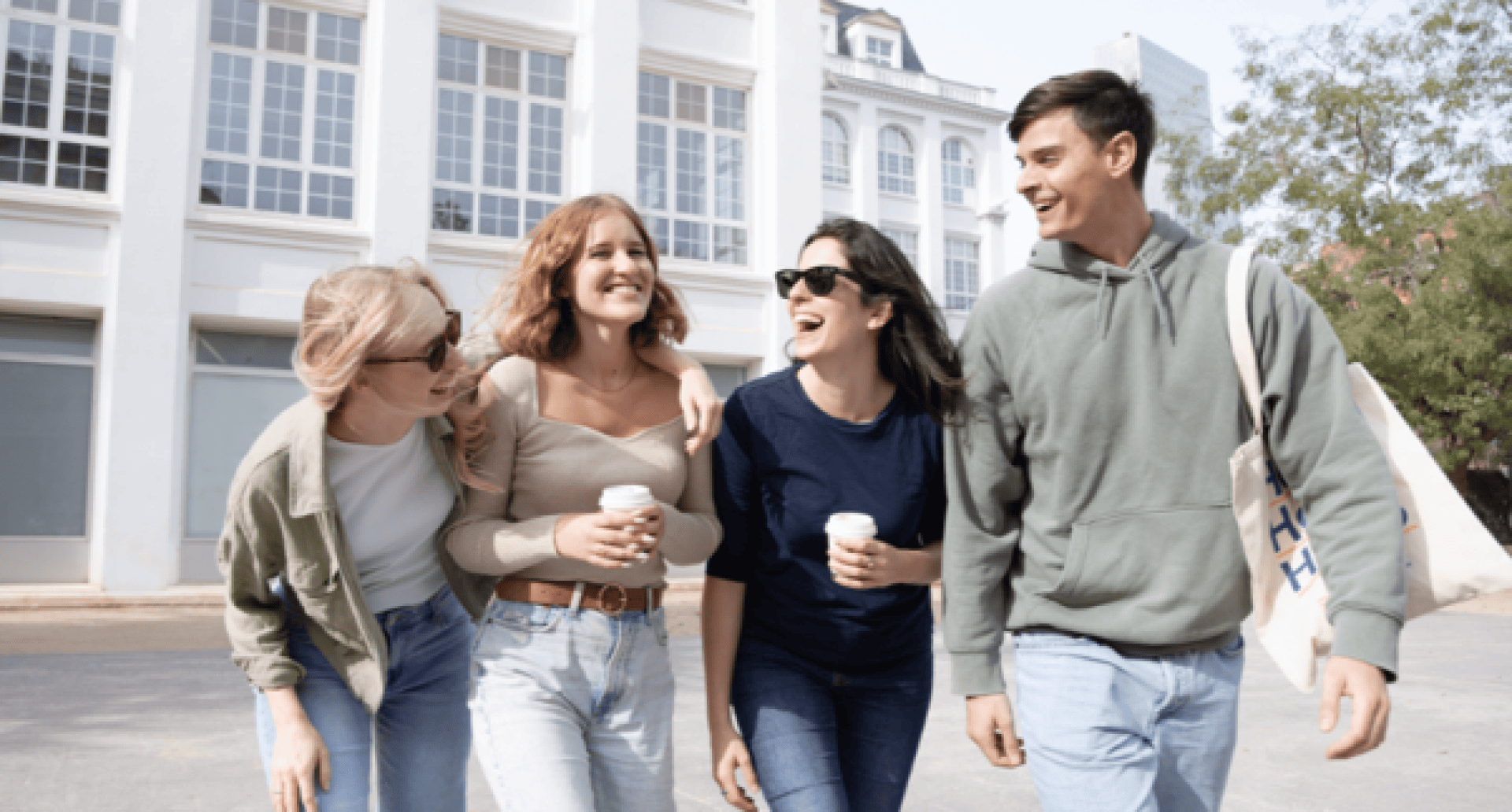 Quatre jeunes adultes se promènent ensemble en plein air, souriants et rieurs. Deux d'entre eux tiennent des tasses de café. Vêtus de tenues décontractées et portant des lunettes de soleil, ils profitent d'une journée ensoleillée devant un bâtiment blanc aux grandes baies vitrées, avec des arbres en arrière-plan.