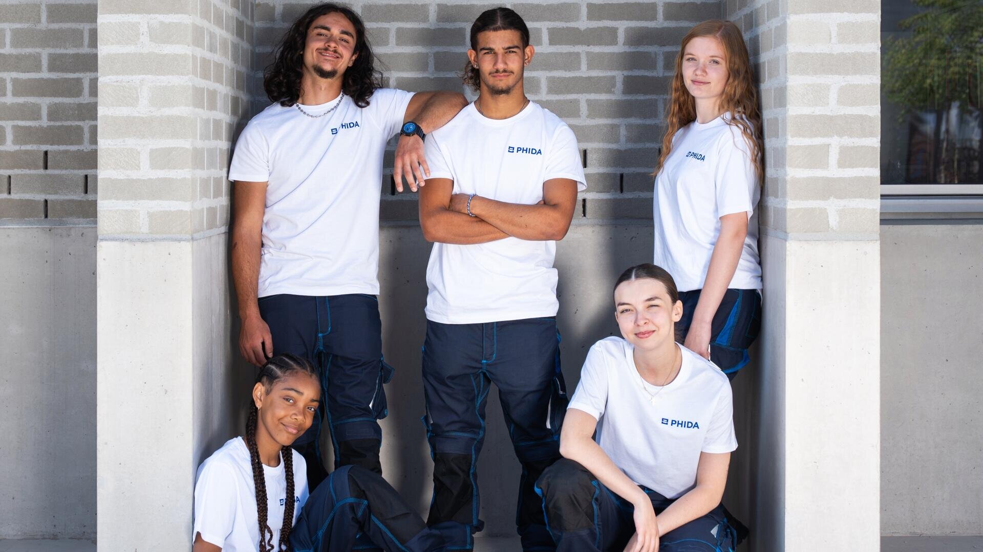 Cinq jeunes adultes vêtus de chemises blanches et de pantalons sombres assortis posent ensemble à l'extérieur, contre un mur de briques claires, en souriant avec assurance. Quatre d'entre eux sont debout et un autre est accroupi devant.