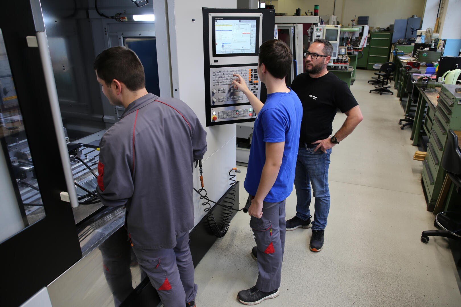 Trois personnes se tiennent près d'une machine à commande numérique dans un atelier. Deux d'entre elles portent des tenues de travail grises ; l'une d'elles manipule le panneau de commande, tandis que la troisième, vêtue d'une chemise noire et d'un jean, observe la scène. On aperçoit à l'arrière-plan du matériel d'atelier et des bureaux.