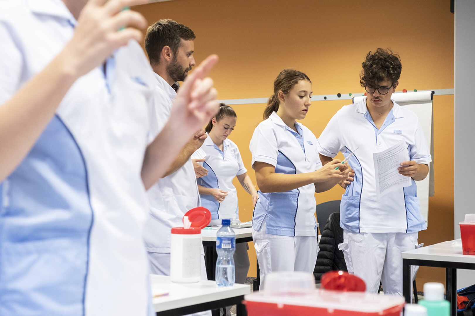 Un groupe d'étudiants infirmiers en uniformes blancs et bleu clair travaillent ensemble dans une salle de classe, certains tenant des fournitures médicales et des papiers, engagés dans une activité pratique.