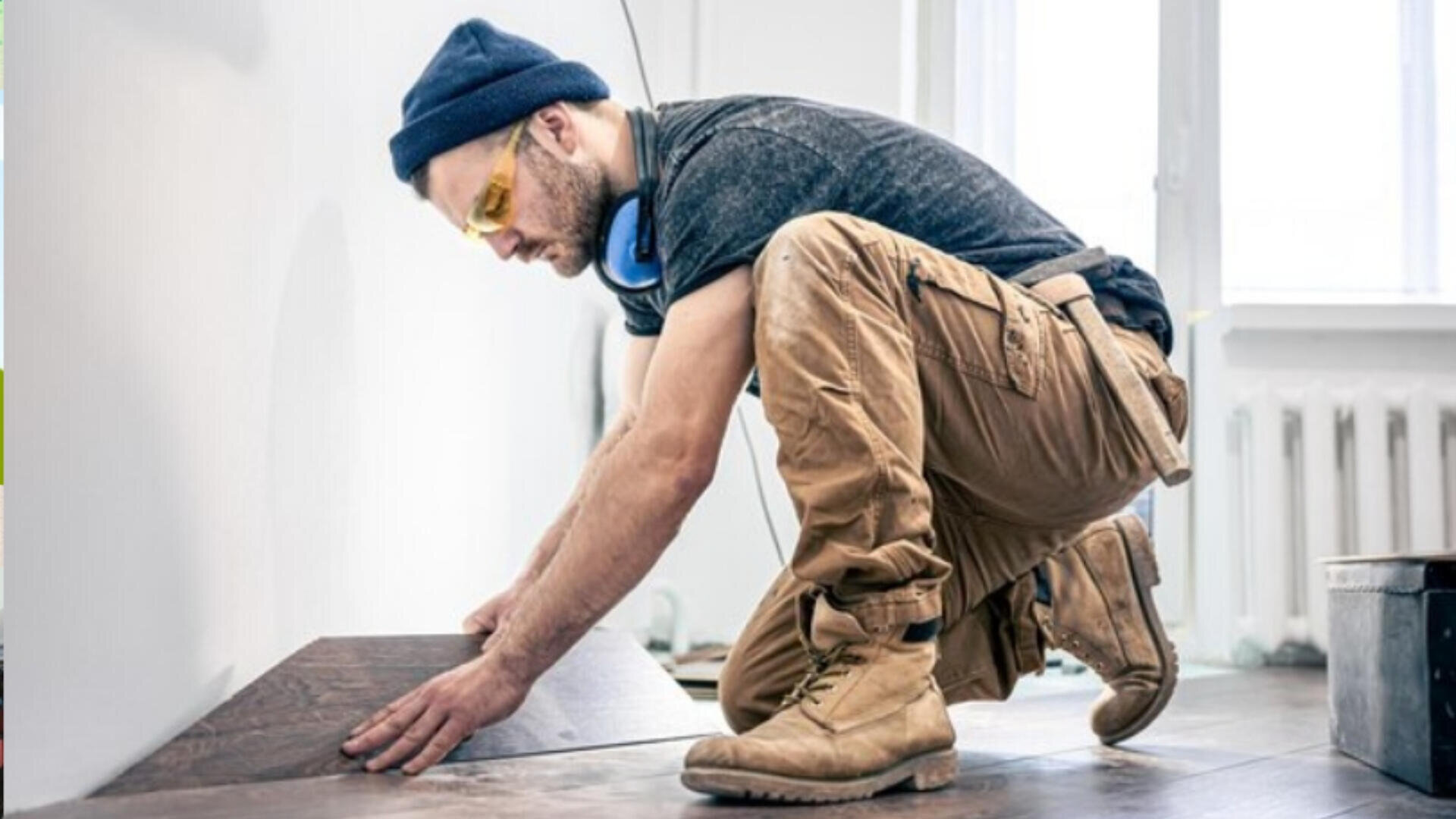 Un homme portant un bonnet, des lunettes de sécurité et des bottes de travail est agenouillé sur le sol, installant soigneusement un parquet en bois foncé le long d'un mur blanc dans une pièce lumineuse.