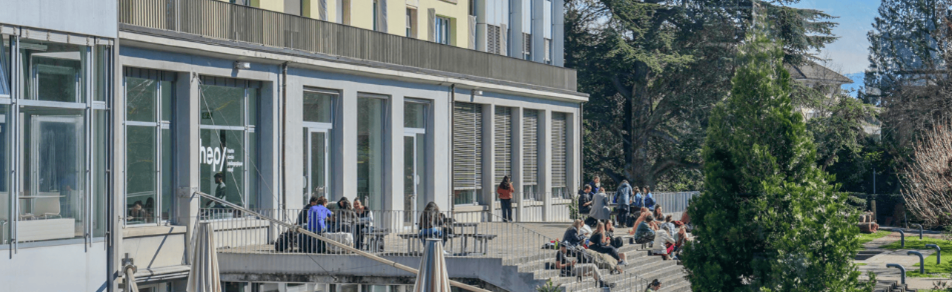 Des étudiants s'assoient et se détendent sur des marches à l'extérieur d'un bâtiment universitaire moderne doté de grandes fenêtres, entouré d'arbres et de verdure lors d'une journée ensoleillée.