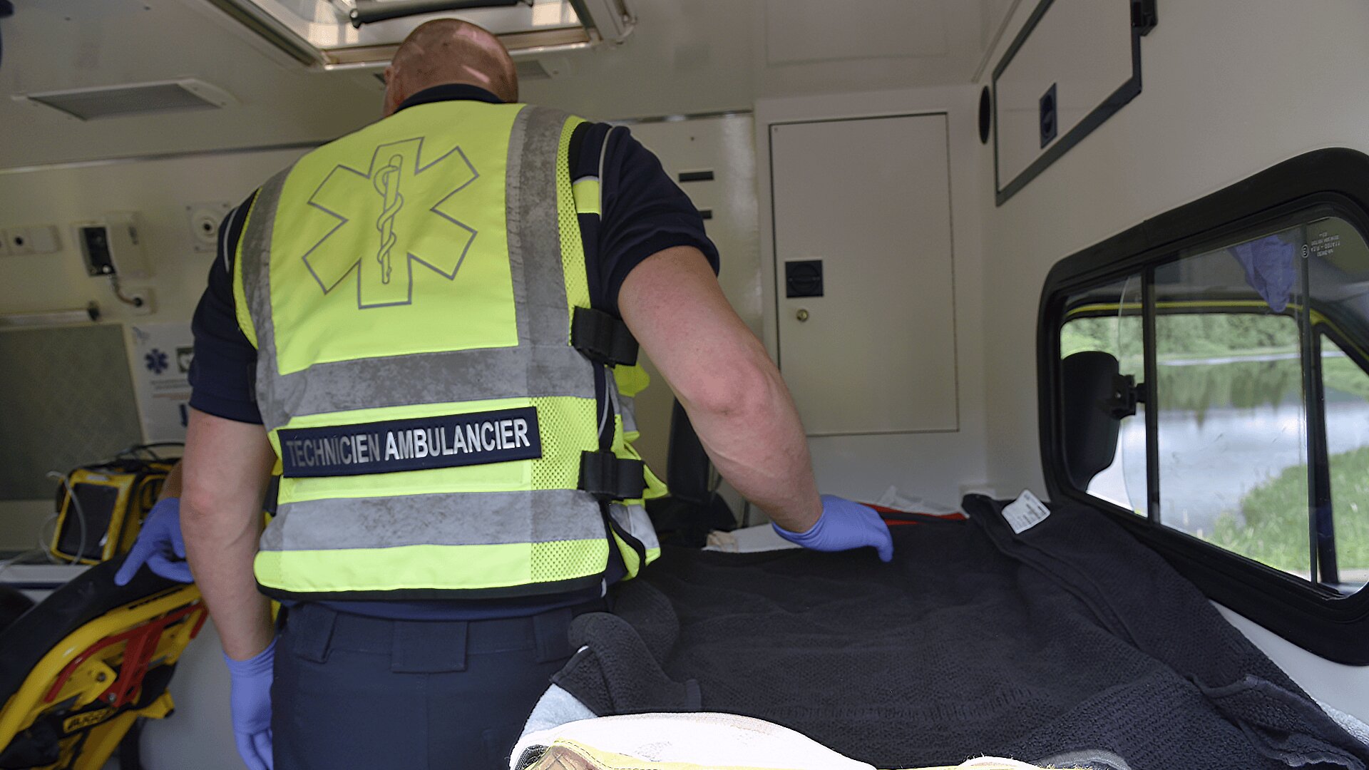 Un secouriste portant un gilet de haute visibilité marqué TECHNICIEN AMBULANCIER se tient à l'intérieur d'une ambulance et s'occupe d'un patient recouvert d'une couverture noire. Des équipements médicaux sont visibles à l'arrière-plan.