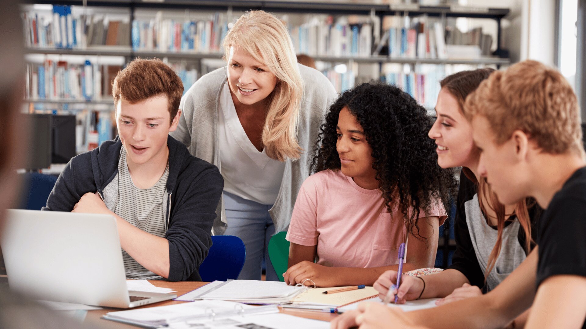 Une enseignante se tient debout et sourit alors qu'elle aide quatre lycéens réunis autour d'un ordinateur portable dans une bibliothèque, avec des livres et des papiers étalés sur la table. Les élèves ont l'air engagés et concentrés sur l'écran.