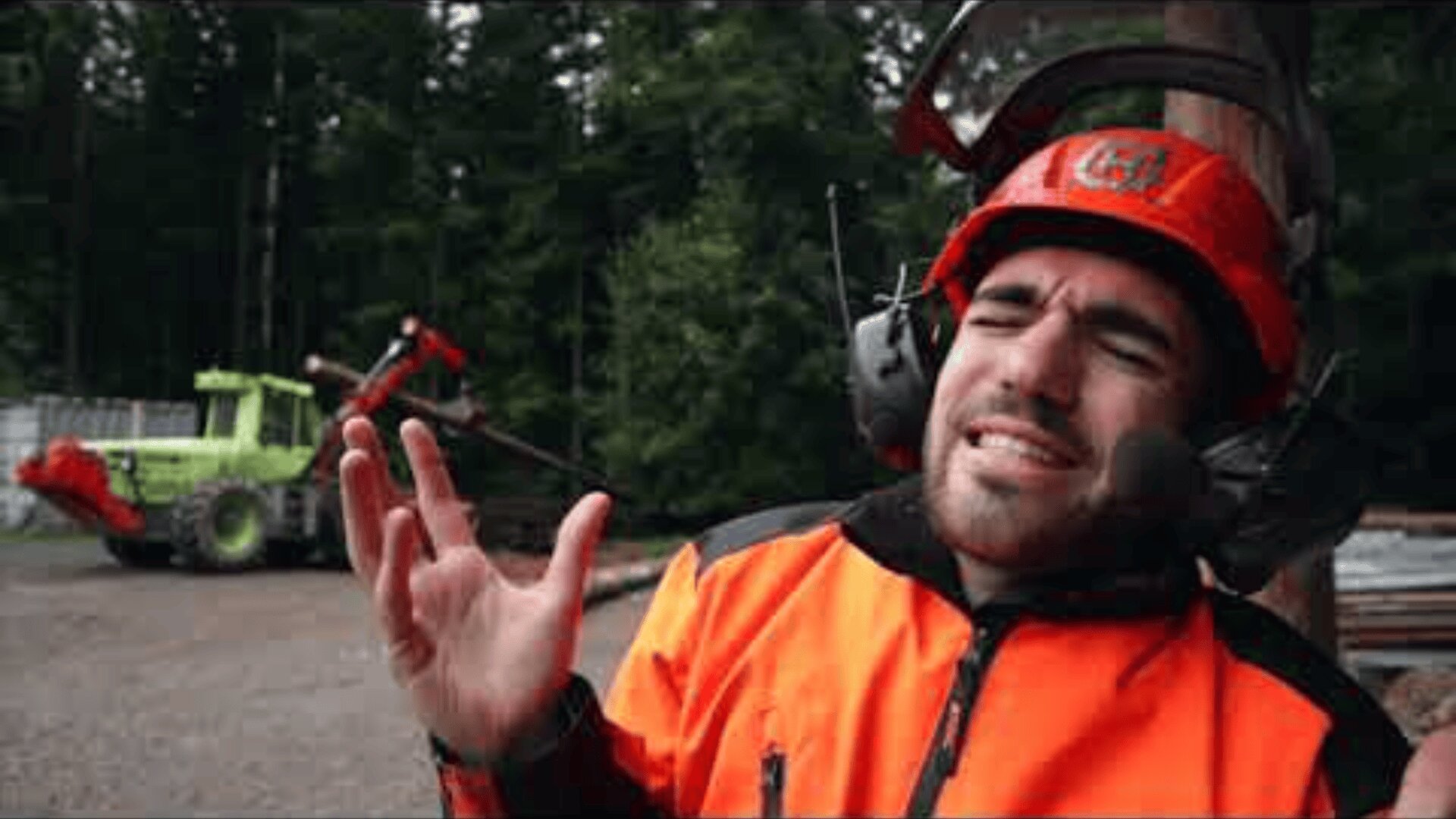 Un homme portant une veste de sécurité et un casque orange fait des gestes expressifs, semblant chanter ou parler avec passion sur un chantier avec des machines et des arbres en arrière-plan.