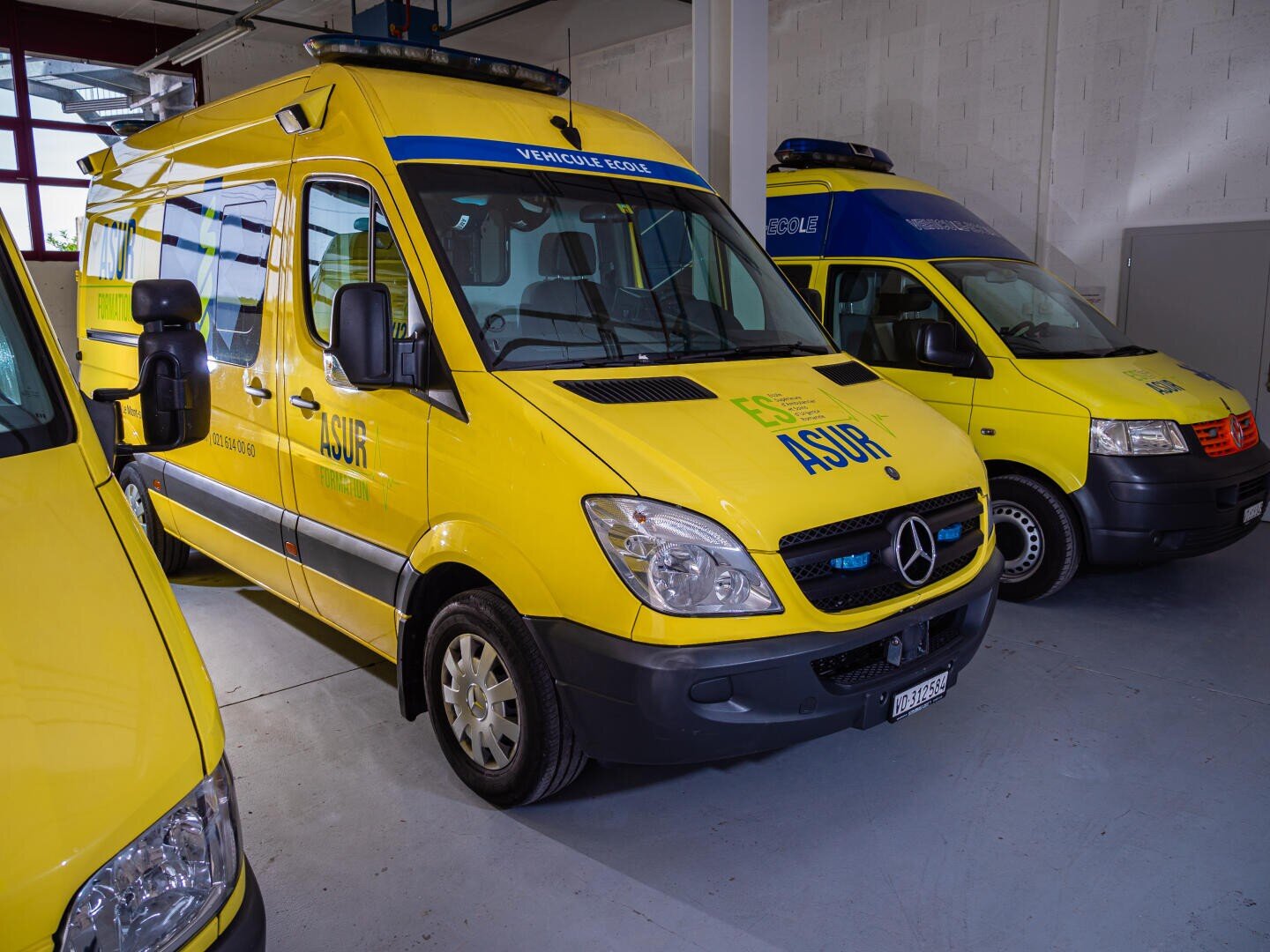 Trois ambulances jaunes sont garées à l'intérieur d'un garage. Les véhicules portent l'inscription ASUR sur le côté et des feux d'urgence bleus sur le dessus. L'ambulance de devant est une Mercedes-Benz.