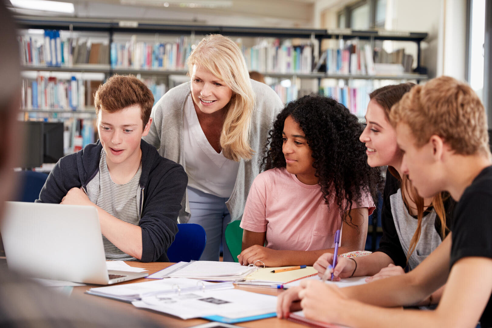Une enseignante souriante se tient derrière quatre lycéens réunis dans une bibliothèque autour d'une table et d'un ordinateur portable, plongés dans leur travail de groupe.