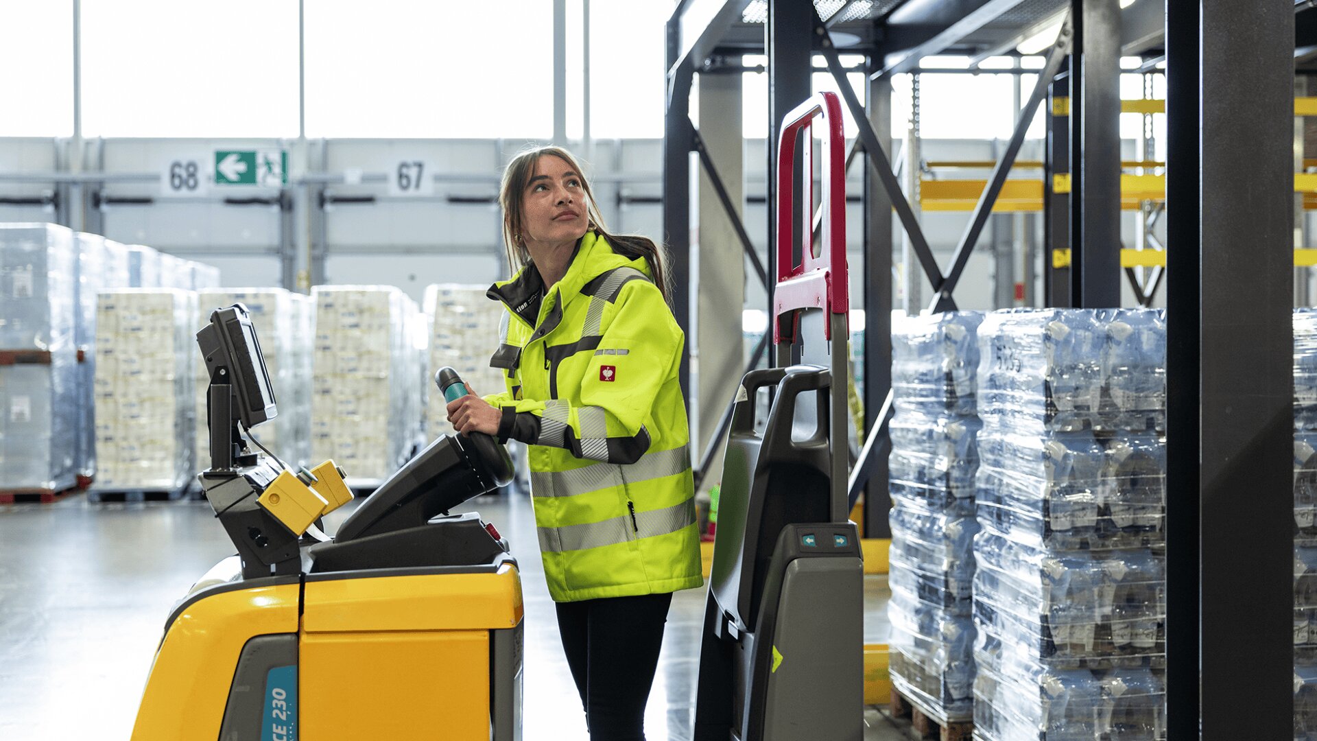 Une femme portant une veste de haute visibilité actionne un transpalette jaune dans un entrepôt, entouré de palettes d'eau en bouteille empilées et de rayonnages industriels.