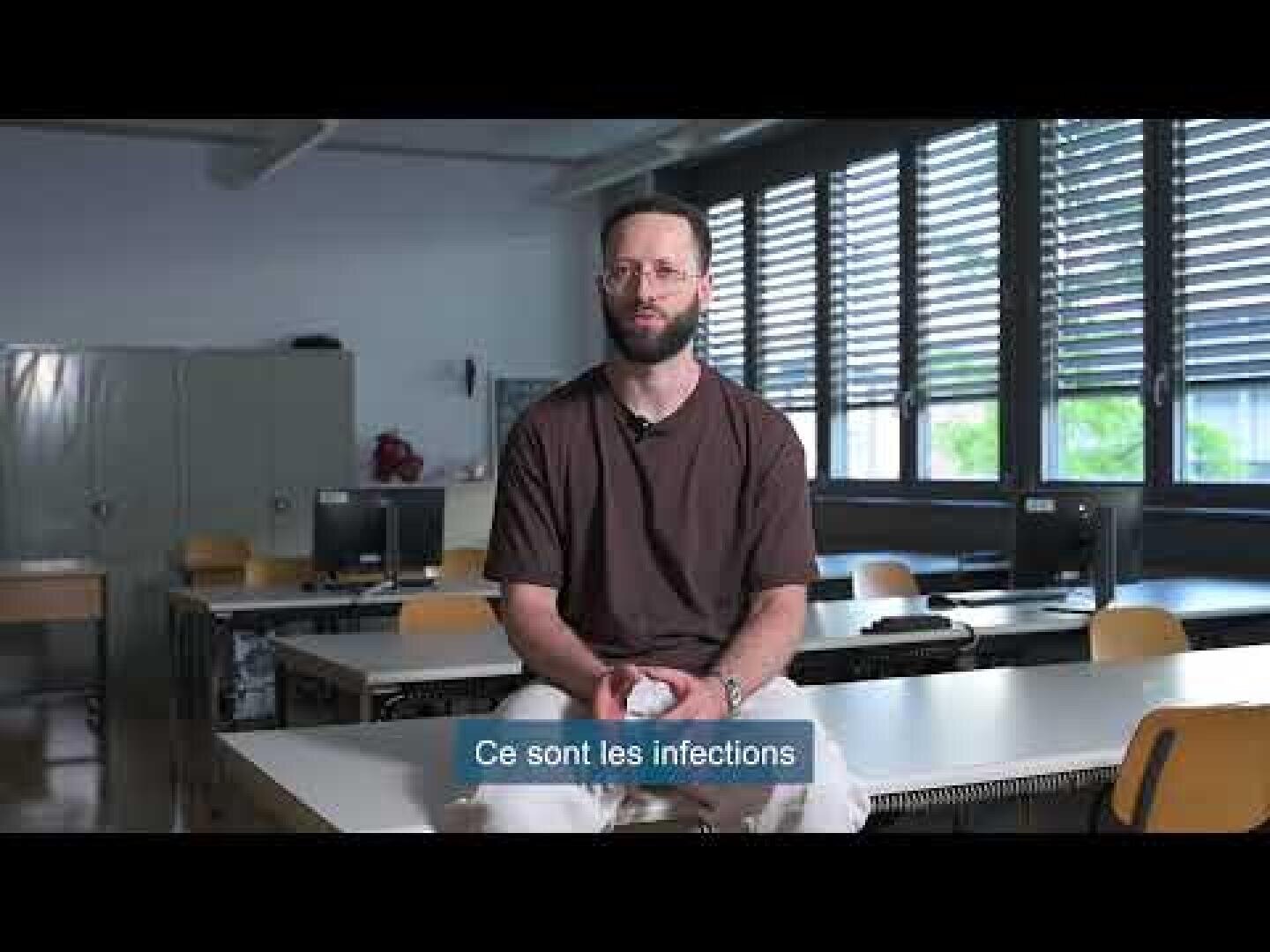 Un homme barbu et portant des lunettes est assis sur une table dans une salle de classe avec des ordinateurs, des armoires et de grandes fenêtres. Les sous-titres français de l'image indiquent : Ce sont les infections.