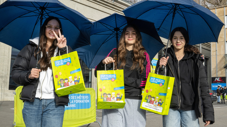 Trois jeunes femmes se tiennent à l'extérieur, tenant des parapluies bleus et des sacs d'événement vert vif, et souriant à l'appareil photo. L'une d'entre elles fait un signe de paix. Elles sont en tenue décontractée, avec un bâtiment et une barrière en arrière-plan.