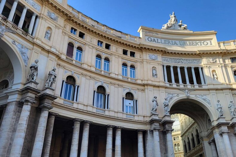 Façade incurvée de la Galleria Umberto I à Naples, en Italie, avec ses grandes fenêtres cintrées, ses colonnes élégantes, ses statues et ses détails architecturaux richement décorés, sur fond de ciel d'un bleu limpide.