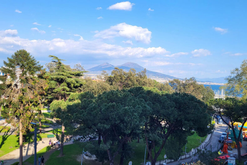 Vue d'un parc aux arbres verdoyants, où des gens se promènent, avec le Vésuve visible en arrière-plan sous un ciel bleu parsemé de nuages.