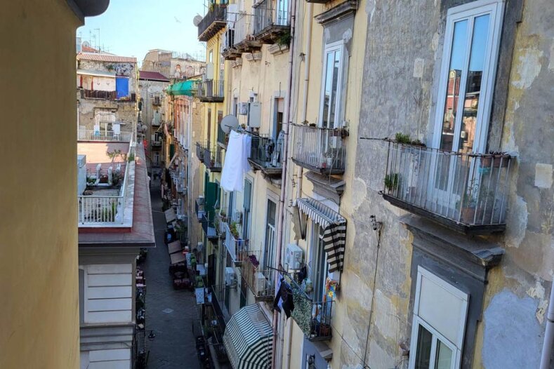 Vue d'une rue étroite bordée d'immeubles anciens et patinés par les intempéries, avec des balcons ornés de plantes et du linge étendu, et quelques passants en contrebas.