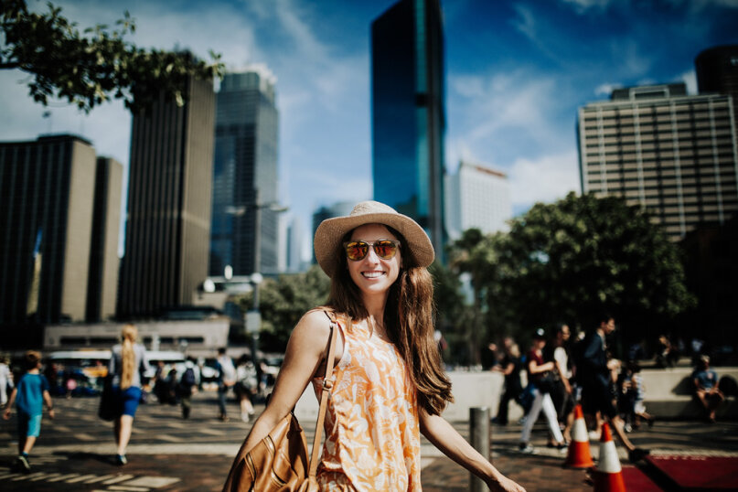 Une femme souriante portant des lunettes de soleil et un chapeau de soleil marche à l'extérieur dans une ville animée, avec de grands bâtiments, des arbres et des gens en arrière-plan par une journée ensoleillée.