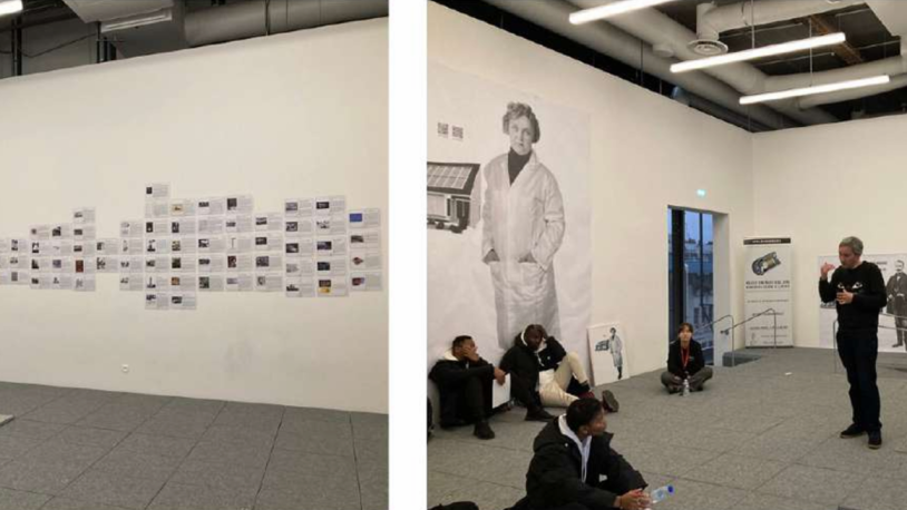 Dans une pièce moderne recouverte d'une moquette grise, des personnes sont assises par terre. Un homme se tient debout et parle. Des documents sont disposés le long d'un mur ; une grande photo en noir et blanc représentant une personne en blouse de laboratoire orne un autre mur.