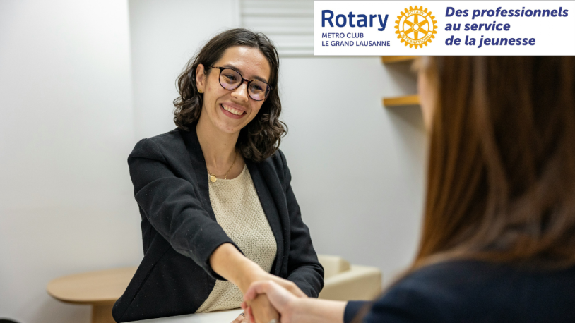 Une femme souriante, vêtue d'un blazer, serre la main d'une autre personne ; le logo et le nom du Rotary Club Le Grand Lausanne sont visibles dans le coin supérieur.
