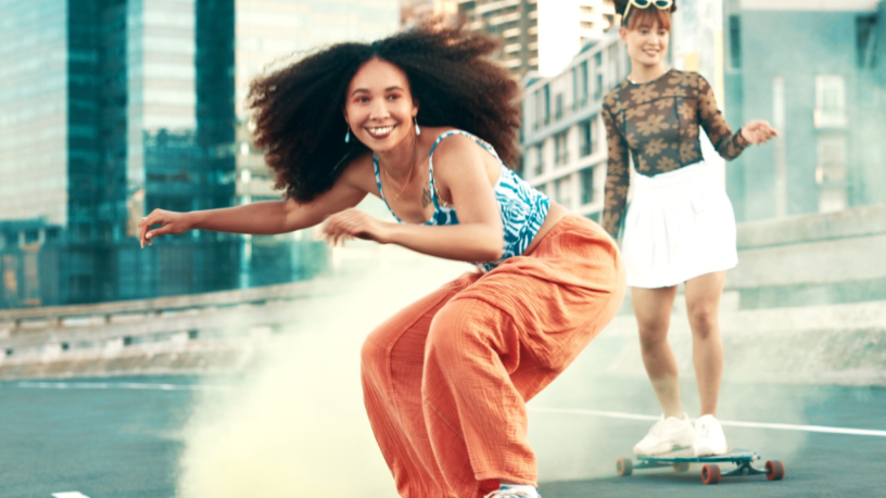 Deux jeunes femmes font du skateboard en plein air dans une ville. La femme de devant sourit, vêtue d'un pantalon orange et d'un débardeur à motifs, avec de la poudre colorée dans l'air. La deuxième femme suit derrière sur un skateboard.