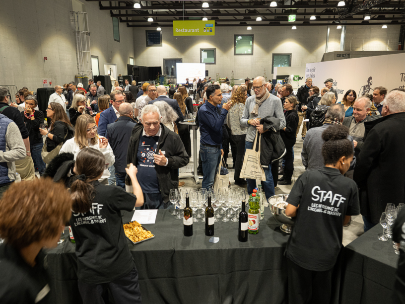 Une foule nombreuse se presse lors d'un événement organisé en salle. Le personnel sert du vin et des amuse-bouches à la table d'accueil, tandis que les invités discutent et dégustent des boissons. Le lieu est bien éclairé, avec de hauts plafonds et une enseigne jaune « Restaurant » visible au-dessus.