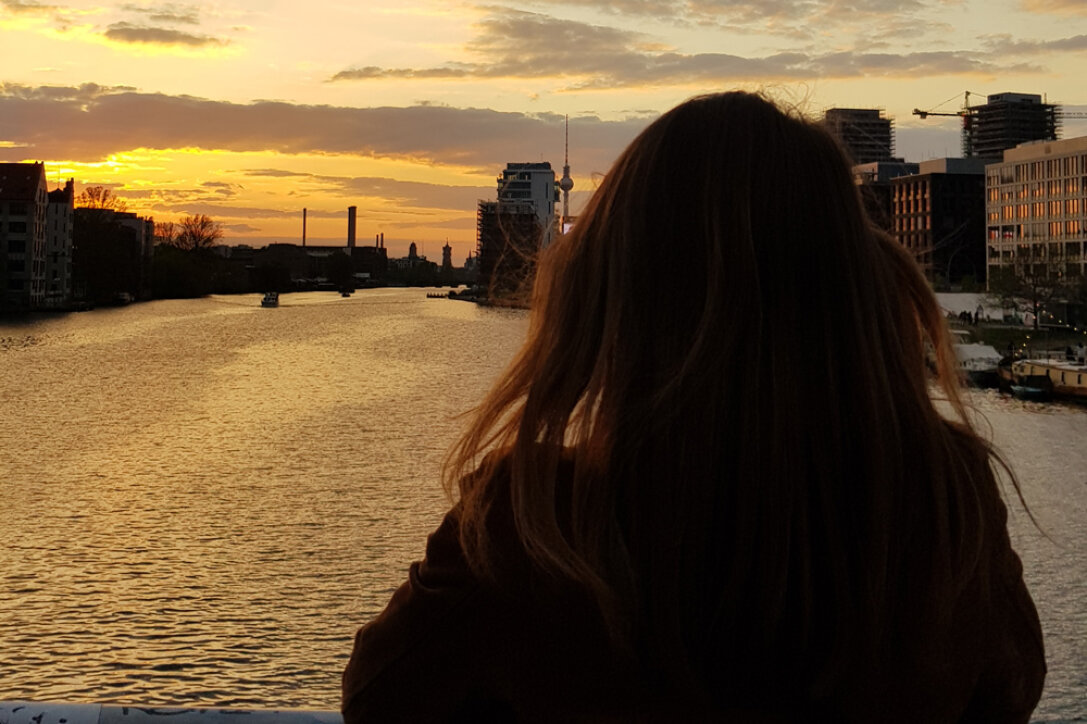 Une personne aux longs cheveux bruns se tient face à une rivière au coucher du soleil, avec des bâtiments urbains et des grues visibles le long de l'eau et un ciel orange en arrière-plan.