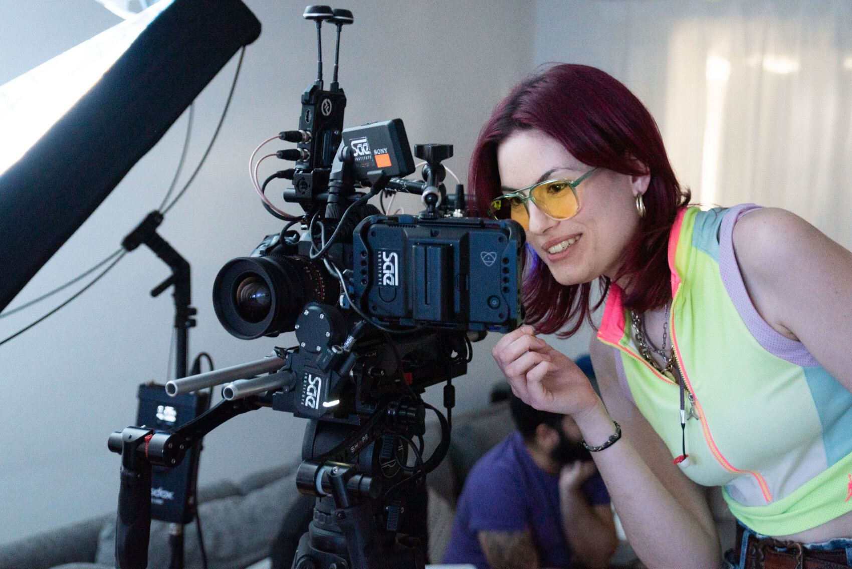 Une femme aux cheveux roux et aux lunettes teintées de jaune sourit tout en réglant une caméra vidéo professionnelle sur un plateau de tournage, vêtue d'un haut sans manches coloré.